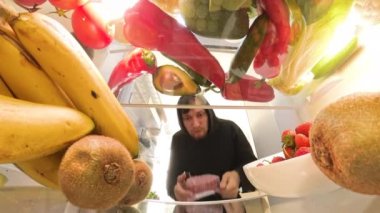 Buzdolabının içinden bir perspektif, kıyma paketini dikkatlice inceleyen bir adamı gösteriyor. Ama en sonunda onu renkli meyve ve sebzelerle çevrili bir şekilde içeride bırakıyor..