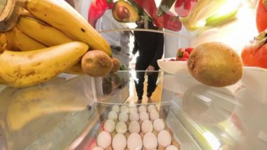 Doli yakınlaştırma efekti, canlı meyve ve sebzelerle çevrili bir yumurtayı almak için buzdolabına uzanan bir adamı parlak ve düzenli bir şekilde yakalar..