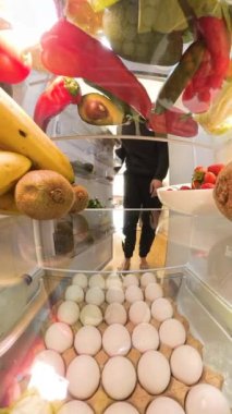 Dikey video. Doli yakınlaştırma efekti, canlı meyve ve sebzelerle çevrili bir yumurtayı almak için buzdolabına uzanan bir adamı parlak ve düzenli bir şekilde yakalar..