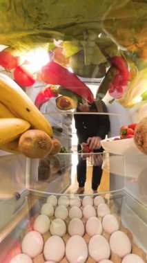 Dikey video. Buzdolabının içinden bir perspektif, canlı meyveler ve renkli ürünlerle çevrili canlı ve düzenli bir şekilde sebze ve yumurta almak isteyen bir adamı gösteriyor..
