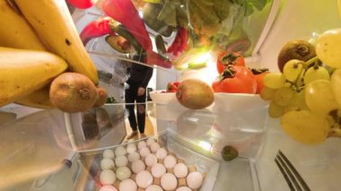 Buzdolabının içinden bir perspektif, canlı meyveler, sebzeler ve parlak ve taze bir ortamda bir tepsi yumurtayla çevrili bir adamın hurmaya uzandığını gösteriyor..