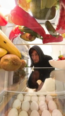 Dikey video. Zoom, buzdolabının içinden bir perspektif, etrafa bakan bir adamı yakalar, ne yiyeceğine karar verirken içeriğini inceler, kamera da onun meraklı bakışlarını takip eder..