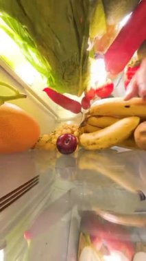 Dikey video. Buzdolabının içinden bir perspektif, bir adamın bir eriği alıp geri koyduğunu gösteriyor. Renkli meyveler, sebzeler ve yumurtalarla çevrili, düzenli ve canlı bir şekilde..