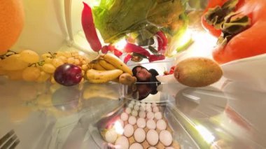 Buzdolabının içinden bir perspektif, canlı ve organize bir sahnede, renkli meyve, sebze ve yumurtalarla çevrili, içeriğini yakından incelemek için öne doğru eğilmiş bir adamı gösteriyor..