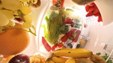 Buzdolabının içinde yakın plan bir görüntü kamerayı canlı meyve ve sebzelerle dolu cam rafların arasından yukarı bakarken gösterir, parlak bir perspektifte dönen bir perspektif yakalar.