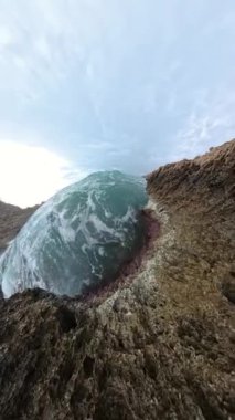Dikey video. Ağır çekimde kayalık bir kayalıklara çarpan yüksek bir dalganın geniş açılı görüntüsü, üzerlerinde dramatik fırtına bulutları bulunan deniz spreylerini ve engebeli dokuları yakalıyor..