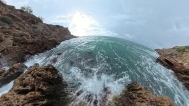 Güçlü bir dalga, kıyı kayalarına çarptığında yavaş çekimde, su damlacıklarını, dramatik bulutlu bir gökyüzünün altında havaya püskürterek,.