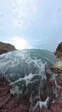 Dikey video. Ağır çekimde bir su altı perspektifi kayalara çarpan bir dalgayı gösteriyor. Kabarcıklar ve girdaplı su detaylı bir şekilde çekiliyor ve dinamik bir deniz manzarası yaratıyor..