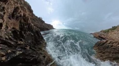 Ağır çekimde, köpüklü su ve bulutlu bir gökyüzünün altındaki çalkantılı deniz ile engebeli kıyı kayaları üzerinde dalgalanan dalgaların dramatik bir genel görünümü..