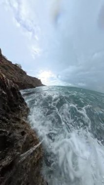 Dikey video. Ağır çekimde, köpüklü su ve bulutlu bir gökyüzünün altındaki çalkantılı deniz ile engebeli kıyı kayaları üzerinde dalgalanan dalgaların dramatik bir genel görünümü..