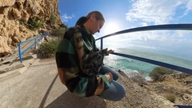 Bir kadın, okyanusa bakan bir tırabzandan kediyi tutar. Güneş ışığı bulutların arasından akar ve dalgalar uzaktan çarpar..