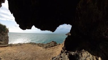 Okyanusa bakan kayalık bir mağaranın içinde, suyun üzerinde parlayan güneş ışığı ve parçalı bulutlu bir gökyüzünün altındaki uzak uçurumların yakın görüntüsü..