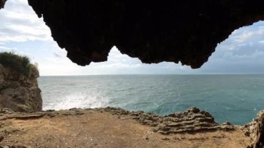 Kapüşonlu gizemli bir adamın, okyanusa bakan bir mağaranın içinde, engebeli kaya dokuları ve arka planda ışıldayan güneş ışığıyla yakın plan..