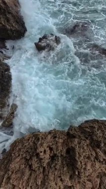 Dikey video. Beyaz köpüklü dalgaların keskin kayalık yüzeylere çarpıp kıvrımlı suyun dokusunu ve gücünü gösteren dramatik bir yavaş çekim..