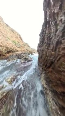 Dikey video. Güçlü bir yavaş çekim dalgası engebeli kıyı kayalarına çarpıyor, su damlacıkları ve köpükler dramatik bir gökyüzü altında havaya uçuyor..