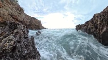 Kayalık uçurumlar arasında dalgaların dalgalandığı, kareyi dolduran köpüklü su ve uçan damlacıklarla dolu, denizin ham enerjisini gösteren yakın çekim bir çekim sahnesi..