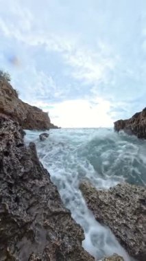 Dikey video. Kayalık uçurumlar arasında dalgaların dalgalandığı, kareyi dolduran köpüklü su ve uçan damlacıklarla dolu, denizin ham enerjisini gösteren yakın çekim bir çekim sahnesi..