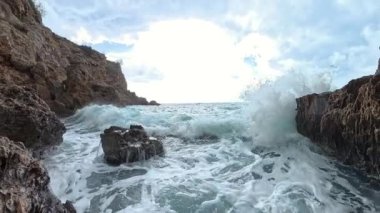 Şiddetli bir dalganın yavaş çekimleri dalgalı kıyı kayalarına çarpıyor, köpüklü su havaya sıçrıyor ve okyanusun ham enerjisi tam ekrana yansıyor..