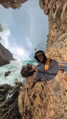 Dikey video. Okyanusun yakınındaki kayalık kayalıklarda duran bir adamın eğri büğrü açılı bir görüntüsü. Dalgalar aşağı düşerken ve güneş ışığı gökyüzüne vururken kamera lensinden püskürtüyor..