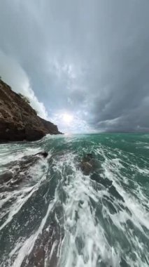 Dikey video. Sarp kayalıklara çarpan dalgaların dramatik yavaş çekim gücü, objektifte su damlacıkları ve okyanus üzerinde fırtına bulutlarını aşan güneş ışığı..