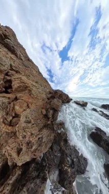 Dikey video. Titrek kayaların arasından geçen okyanus dalgalarının dinamik ve yavaş çekim perspektifi parlak gökyüzünü ve çalkantılı suyu canlı detaylarıyla vurguluyor..