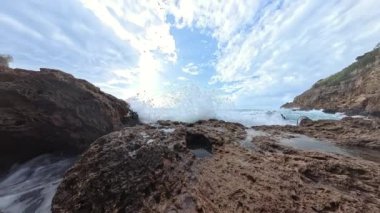 Sivri kayaların üzerinde dalgalanan köpüklü dalgaların yavaş çekim görüntüsü, yükselen kayalıklar ve dağınık bulutların arasından süzülen güneş ışığıyla parlayan bir gökyüzü tarafından çerçevelenmiş..