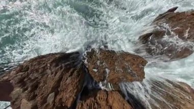 Sarsıntılı kayalara çarpan dalganın patlama kuvvetini gösteren dramatik bir yukarıdan çekim, kamera aşağıdaki dalgalanan sudan kaçınmak için yükseliyor gibi görünüyor..