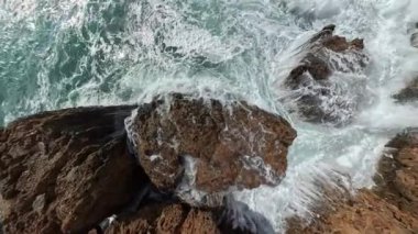 Keskin kıyı kayaları etrafında dönen ve köpüren dalgaların hava perspektifi. Kamera, çalkantılı suyun güçlü etkisinden uzaklaşıyor..