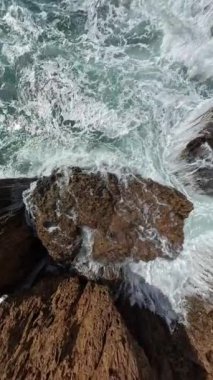 Dikey video. Keskin kıyı kayaları etrafında dönen ve köpüren dalgaların hava perspektifi. Kamera, çalkantılı suyun güçlü etkisinden uzaklaşıyor..