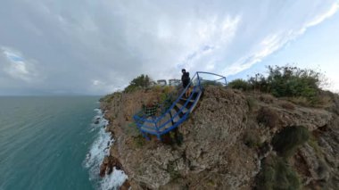 Mavi bir merdivende duran bir adam ve bir kadının geniş açılı görüntüsü sarp bir uçuruma tünemiş, dramatik bir gökyüzünün altındaki engin turkuaz denize bakıyor..