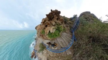 Kıvrımlı bir merdiven, yeşilliklerle çevrili engebeli bir uçurumdan aşağı süzülüyor. Denize doğru inen sarı bir sırt çantası olan bir adam, kayalık dokularla çevrelenmiş..