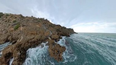 Kayalık uçurumların ve bulutlu bir gökyüzünün altındaki okyanusun geniş açılı görüntüsü kayalıklardaki insanları ve aşağıdaki dalgaların çarptığı uçurum boyunca uzanan dar bir yolu gösteriyor..