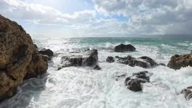 Güçlü dalgalar, dalgalı kıyı kayalarının üzerine çarpıyor, köpüklü su engebeli araziye yayılıyor dramatik bulutlarla dolu güneşli bir gökyüzü altında..