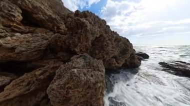 Güçlü dalgaların panoramik görüntüsü parlak bir güneş ve dramatik bulutlar altında sivri kayaların üzerinde köpüren deniz manzarası, deniz manzarasının vahşi güzelliğini yakalıyor..