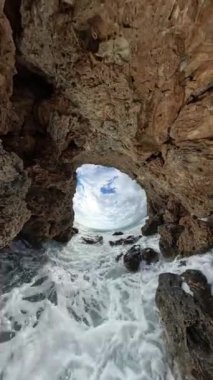 Dikey video. Okyanusu çevreleyen doğal bir kaya kemerinin içinden bakın, aşağıdaki kayalara çarpan dalgalar ve açıklığın ötesinde dramatik bulutlu bir gökyüzü.