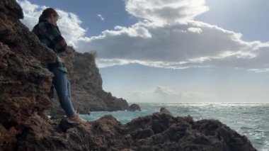 Bir kadın engebeli kıyı kayalarında duruyor, okyanusa bakıyor, güneş ışığı parlak gökyüzündeki dramatik bulutların arasından süzülüyor..