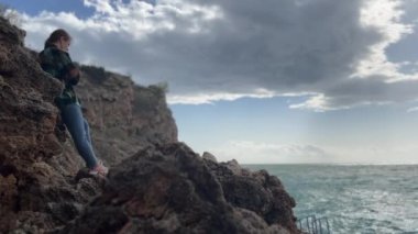 Yalnız bir kadın sivri kayalara yaslanır. Yüzü okyanusa bakarken yansıtır. Rüzgarlı gökyüzü dramatik bulutlarla doludur..