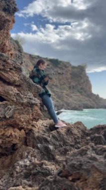 Dikey video. Ekose bir cekete sarılmış bir kadın, dalgalı kıyı kayaları üzerinde duruyor, karamsar, rüzgarlı bir gökyüzünün altında düşünceli ve yalnız bir ifadeyle okyanusa bakıyor..