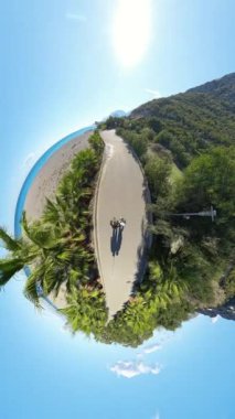 Dikey video. Yemyeşil ve kayalıklarla çevrili kıvrımlı bir patikayı gösteren küçük bir gezegen manzarası, canlı mavi bir gökyüzünün altında kumsalda yürüyen iki kişi..