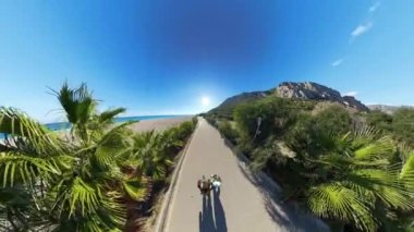 Dönen bir kameranın hava görüntüsü, iki insanı, parlak mavi bir gökyüzünün altında palmiye ağaçları, dağlar ve denizle kaplı manzaralı bir sahil yolunda yürürken yakalıyor..