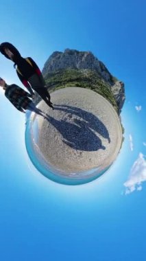 Dikey video. Turkuaz su, yeşil uçurumlar ve canlı mavi gökyüzü ile çevrili, çakıllı bir sahilde yürüyen bir çifti içeren dönen küçük bir gezegen etkisi..