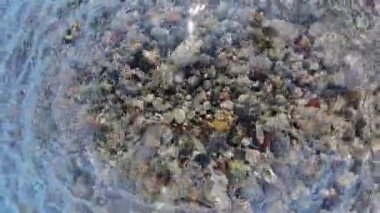 Sahilde pürüzsüz, çok renkli çakıl taşları üzerinde akan, parlak mavi gökyüzünün altında dokuları ve güneş ışığını yansıtan yakın plan bir su..