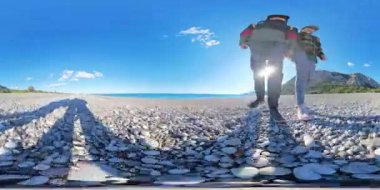 Çakıl taşlı bir plajda yürürken, parlak mavi gökyüzünde parlayan güneşin altında uzun gölgeler bırakan bir çiftin 360 derecelik kusursuz panoraması..