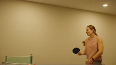 Neşeli bir kadın pinpon topuna vururken dans eder, masa tenisinin yakınındaki kapalı alanda elinde bir kürek tutar, neşeli ve enerjik bir anın tadını çıkarır..