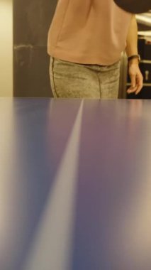 Dikey video. Kapalı alandaki mavi bir masada masa tenisi topuna vuran bir kişinin yakın açılı çekimi sıradan bir oyun düzeninde aksiyon ve hareketi vurguluyor..
