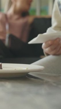 Dikey video. Küçük bir parça çikolatalı kek mermer bir masada beyaz bir tabakta oturur, arka planda buzlu içecekler, rahat ve rahat bir yemek atmosferi yaratır..