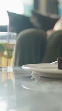 Dikey video. Çikolatalı kek parçasının yakın plan resmi beyaz bir tabakta, arka planda naneli buzlu bir içecek mermer bir masanın üzerinde, ışığı yakalıyor ve yemek sahnesini canlandırıyor..