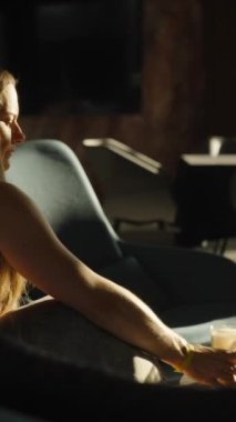 Dikey video. Bir kadın karanlık, şık bir odada otururken latte içmekten zevk alır, batan güneşin altın ışığıyla aydınlanır, pencereden süzülür, huzur verici bir hava yaratır..