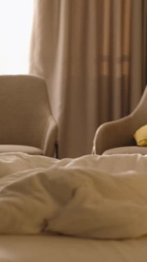 Dikey video. Mobilyalarla çerçevelenmiş, yatakta uzanan bir kadının arka ışıklandırılmış görüntüsü. Sıcak güneş ışığı, modern, rahat bir yatak odasını yumuşak, sessiz tonlarla ve sabah huzuruyla dolduruyor..