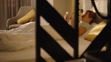 Beyaz bir yorganın altında, sarı aksanlı ve modern yatak odasıyla çevrili, sabah güneşinde akıllı telefonunu kullanan bir kadın..